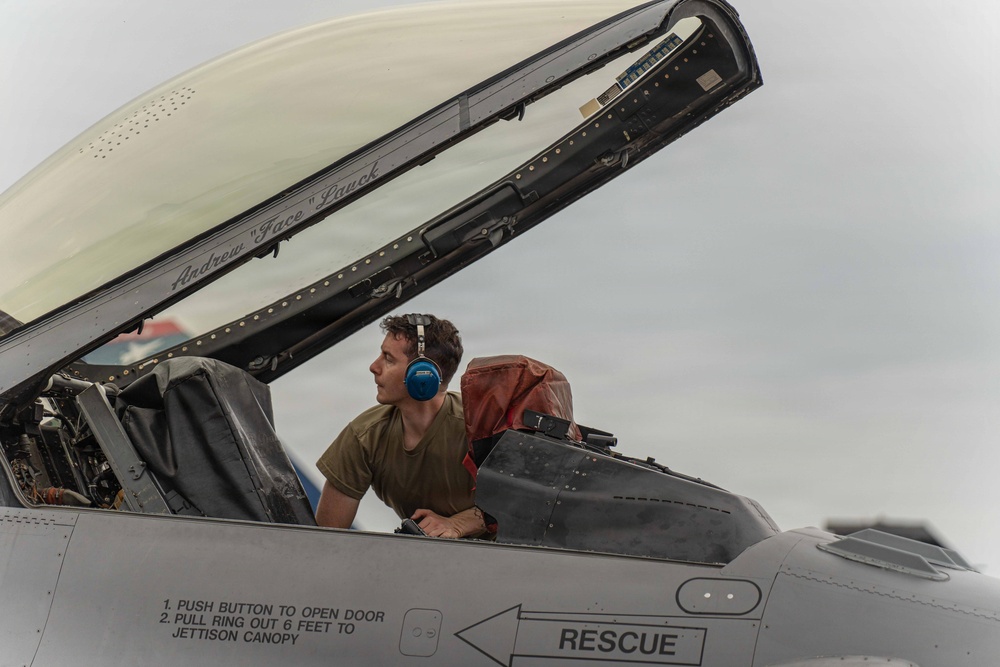 A ground crew technician wearing blue hearing protection is inspecting or servicing the open cockpit of a military fighter jet. The canopy is raised, revealing the ejection seat with a red survival kit, and the aircraft fuselage displays standard rescue markings and canopy jettison instructions. The pilot's name 'Andrew "Face" Lauck' is inscribed on the canopy rail.