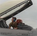 A ground crew technician wearing blue hearing protection is inspecting or servicing the open cockpit of a military fighter jet. The canopy is raised, revealing the ejection seat with a red survival kit, and the aircraft fuselage displays standard rescue markings and canopy jettison instructions. The pilot's name 'Andrew "Face" Lauck' is inscribed on the canopy rail.