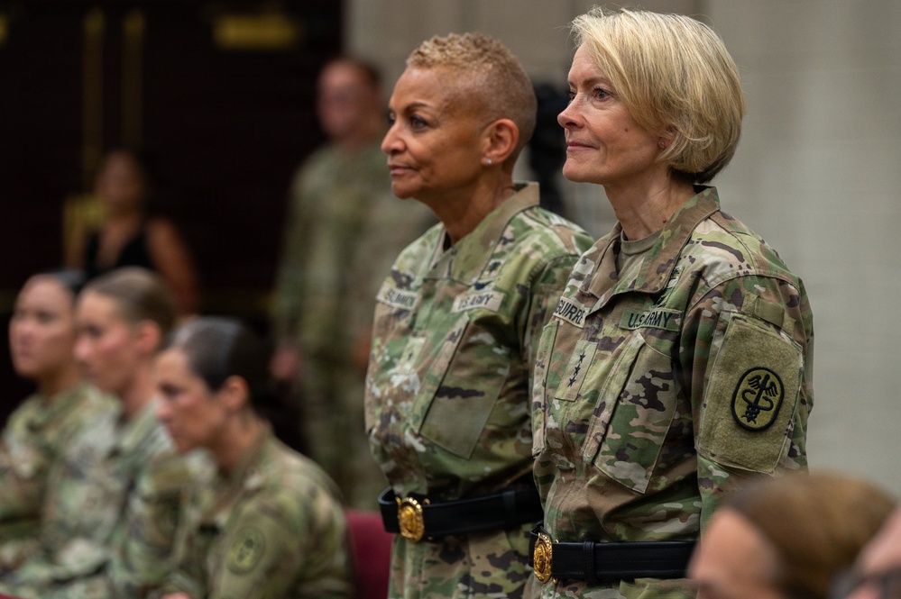 Two U.S. Army female officers stand at attention in the foreground wearing Operational Camouflage Pattern (OCP) uniforms. The officer on the right displays a Medical Corps branch insignia patch and four-star rank indicators, suggesting senior leadership. Several other uniformed personnel are seated in the background in what appears to be a formal military ceremony or event.
