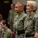 Two U.S. Army female officers stand at attention in the foreground wearing Operational Camouflage Pattern (OCP) uniforms. The officer on the right displays a Medical Corps branch insignia patch and four-star rank indicators, suggesting senior leadership. Several other uniformed personnel are seated in the background in what appears to be a formal military ceremony or event.
