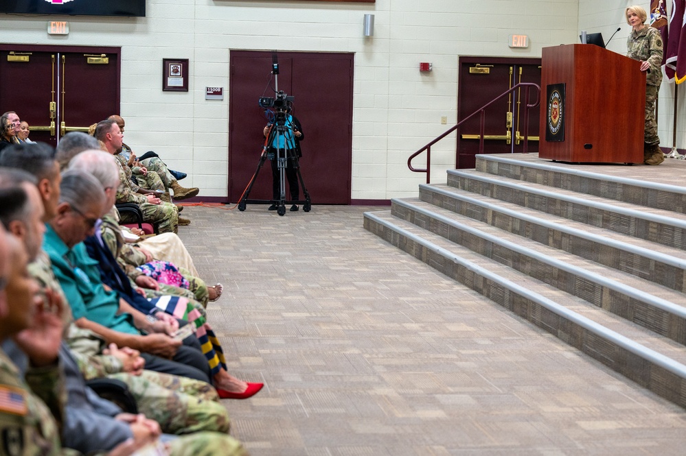 A military ceremony or official event is taking place in what appears to be a formal hall or auditorium. A person in military camouflage uniform is speaking at a wooden podium on a raised stage, addressing a seated audience that includes both military personnel in uniform and civilians. A videographer with a camera on a tripod is positioned in the center of the room to document the event.