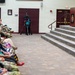 A military ceremony or official event is taking place in what appears to be a formal hall or auditorium. A person in military camouflage uniform is speaking at a wooden podium on a raised stage, addressing a seated audience that includes both military personnel in uniform and civilians. A videographer with a camera on a tripod is positioned in the center of the room to document the event.
