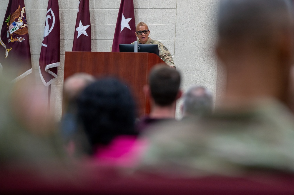 A female military officer in camouflage uniform is speaking at a wooden podium during a formal military briefing or ceremony. She is wearing glasses and addressing an audience of seated military personnel and civilians. Behind her are maroon/burgundy flags bearing white stars, indicating a general officer's presence or command.