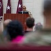 A female military officer in camouflage uniform is speaking at a wooden podium during a formal military briefing or ceremony. She is wearing glasses and addressing an audience of seated military personnel and civilians. Behind her are maroon/burgundy flags bearing white stars, indicating a general officer's presence or command.