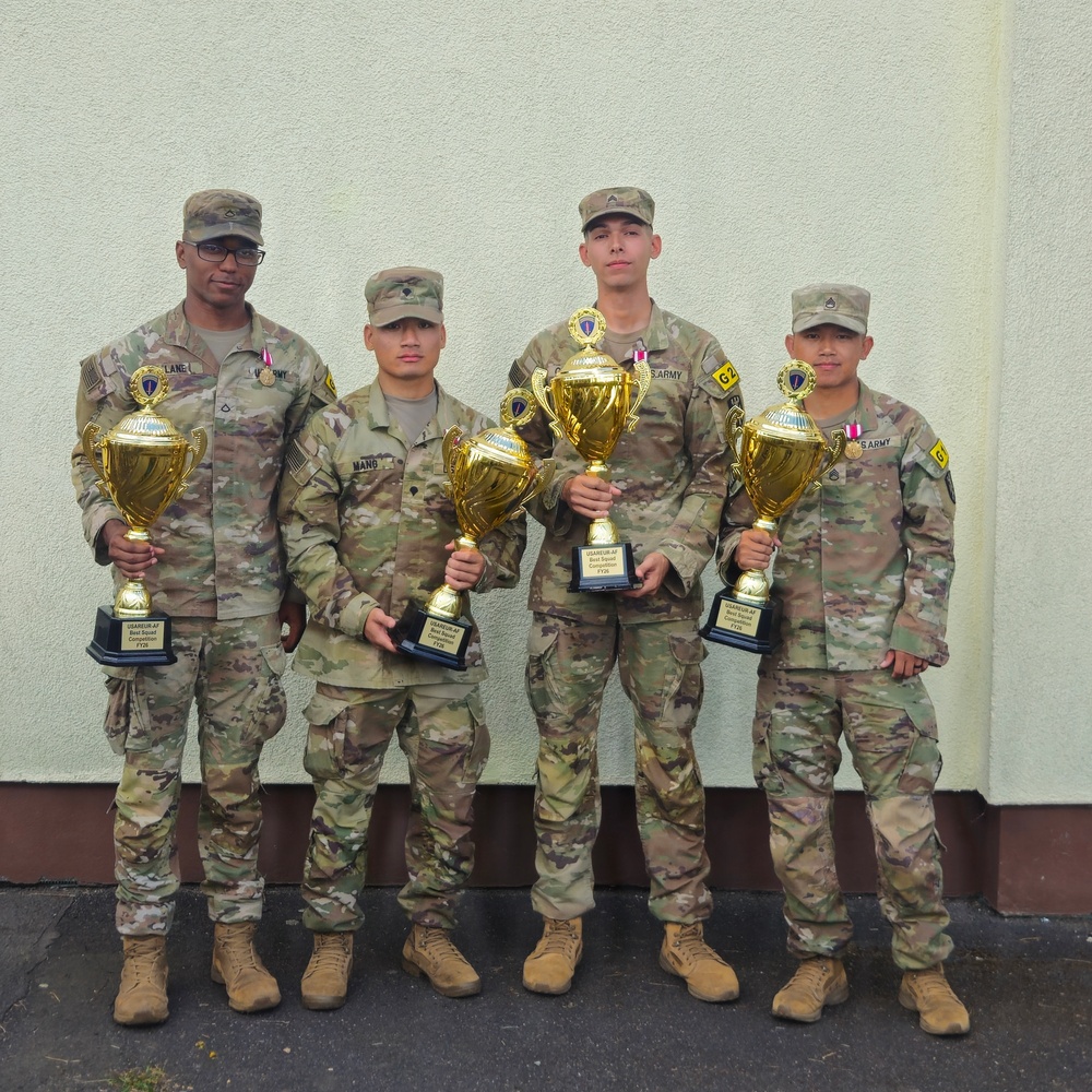 Four U.S. Army soldiers in OCP (Operational Camouflage Pattern) uniforms pose together holding gold trophy cups. The trophies appear to be from a USAREUR-AF competition, with visible text referencing 'Best Squad Competition FY26'. The soldiers are standing in front of a light-colored building exterior.