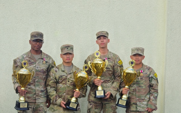 Four U.S. Army soldiers in OCP (Operational Camouflage Pattern) uniforms pose together holding gold trophy cups. The trophies appear to be from a USAREUR-AF competition, with visible text referencing 'Best Squad Competition FY26'. The soldiers are standing in front of a light-colored building exterior.