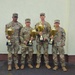 Four U.S. Army soldiers in OCP (Operational Camouflage Pattern) uniforms pose together holding gold trophy cups. The trophies appear to be from a USAREUR-AF competition, with visible text referencing 'Best Squad Competition FY26'. The soldiers are standing in front of a light-colored building exterior.