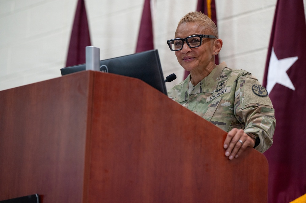 A U.S. Army officer in OCP (Operational Camouflage Pattern) uniform stands at a wooden podium, appearing to address an audience. Behind her are maroon/burgundy military guidon flags, one of which displays a white star indicating a general officer's presence or command. A Dell monitor is visible on the podium.