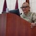 A U.S. Army officer in OCP (Operational Camouflage Pattern) uniform stands at a wooden podium, appearing to address an audience. Behind her are maroon/burgundy military guidon flags, one of which displays a white star indicating a general officer's presence or command. A Dell monitor is visible on the podium.
