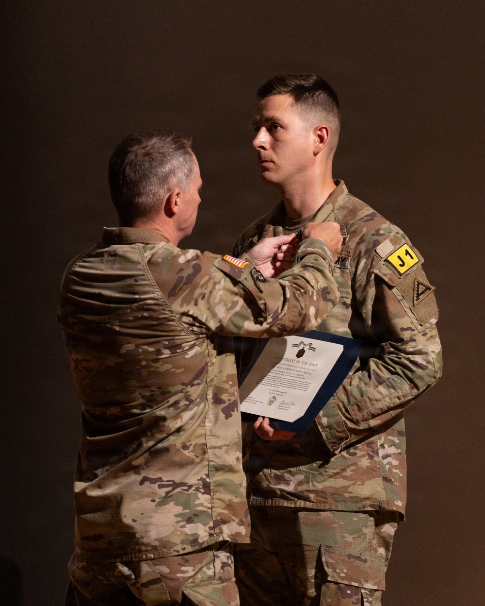 Two U.S. Army soldiers in MultiCam/OCP camouflage uniforms are participating in a military awards ceremony. An older, senior soldier is pinning a medal onto the chest of a younger soldier, who is holding an official Department of the Army certificate. The ceremony appears to be a formal recognition event, likely awarding an Army Commendation Medal based on the visible certificate.