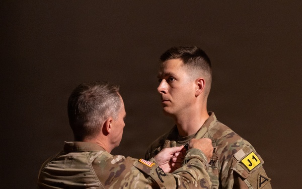 Two U.S. Army soldiers in MultiCam/OCP camouflage uniforms are participating in a military awards ceremony. An older, senior soldier is pinning a medal onto the chest of a younger soldier, who is holding an official Department of the Army certificate. The ceremony appears to be a formal recognition event, likely awarding an Army Commendation Medal based on the visible certificate.