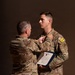 Two U.S. Army soldiers in MultiCam/OCP camouflage uniforms are participating in a military awards ceremony. An older, senior soldier is pinning a medal onto the chest of a younger soldier, who is holding an official Department of the Army certificate. The ceremony appears to be a formal recognition event, likely awarding an Army Commendation Medal based on the visible certificate.