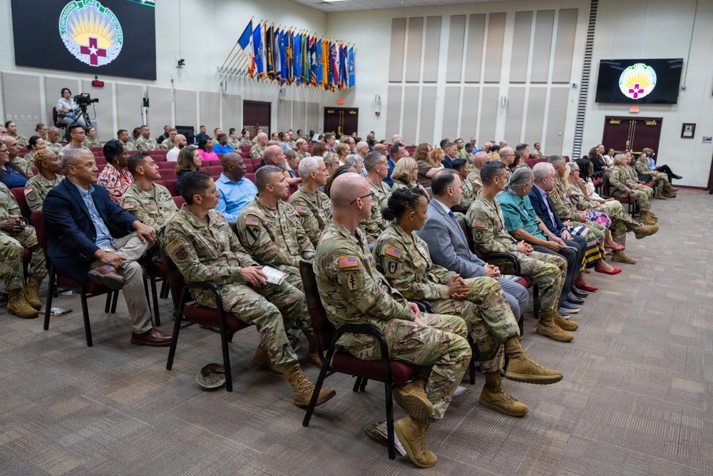 A formal military ceremony or briefing is taking place in an auditorium-style room, with a large audience composed primarily of U.S. Army soldiers in camouflage uniforms (ACUs/OCP pattern) along with some civilians in business attire. The room features multiple military unit flags/guidons displayed along the back wall and an organizational emblem visible on both a large black sign and a monitor screen. The audience is seated in rows of maroon chairs, all facing forward attentively.