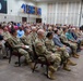 A formal military ceremony or briefing is taking place in an auditorium-style room, with a large audience composed primarily of U.S. Army soldiers in camouflage uniforms (ACUs/OCP pattern) along with some civilians in business attire. The room features multiple military unit flags/guidons displayed along the back wall and an organizational emblem visible on both a large black sign and a monitor screen. The audience is seated in rows of maroon chairs, all facing forward attentively.