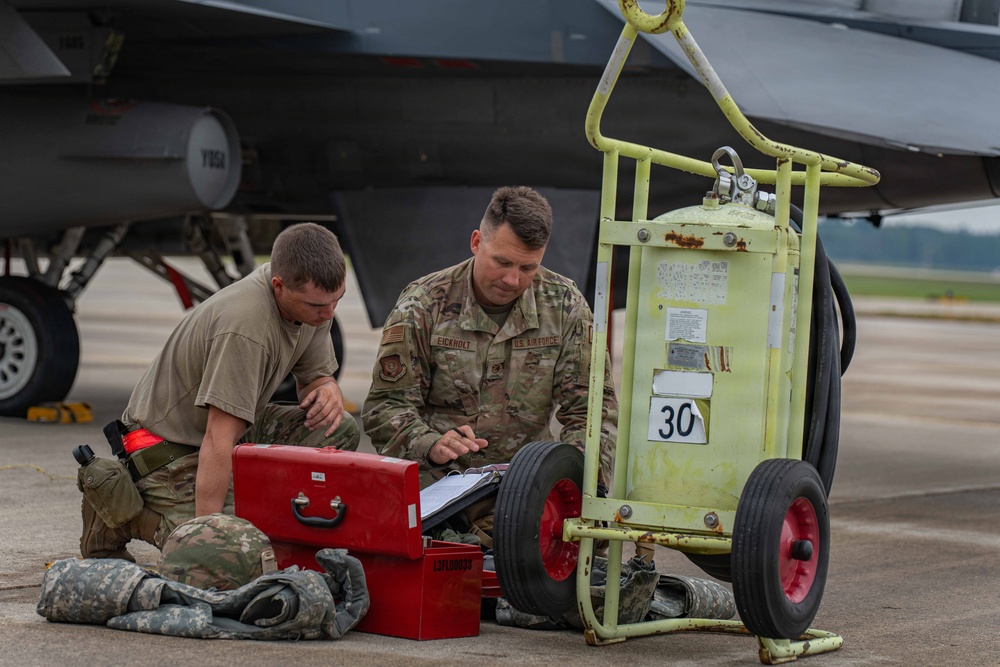 Two U.S. Air Force personnel are kneeling on a flight line tarmac reviewing documents on a clipboard next to a red tool case and a yellow ground support equipment cart numbered '30'. The uniformed airman on the right has a name tape reading 'EICKHOLT' and U.S. Air Force insignia visible. A military aircraft, likely an F-16 or similar fighter jet, is visible in the background with what appears to be a missile or munition mounted under the wing.
