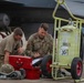 Two U.S. Air Force personnel are kneeling on a flight line tarmac reviewing documents on a clipboard next to a red tool case and a yellow ground support equipment cart numbered '30'. The uniformed airman on the right has a name tape reading 'EICKHOLT' and U.S. Air Force insignia visible. A military aircraft, likely an F-16 or similar fighter jet, is visible in the background with what appears to be a missile or munition mounted under the wing.