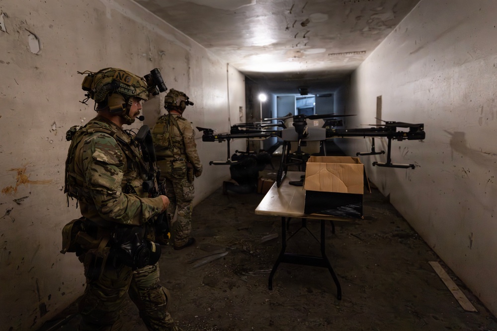 Two military personnel in full combat gear are positioned inside a narrow, dimly lit corridor or tunnel alongside multiple large multi-rotor drones staged on or near a folding table. The soldiers are wearing multicam uniforms, tactical helmets with night-vision mounts, and communication headsets. The setting appears to be a training or operational environment involving unmanned aerial systems (UAS).
