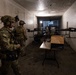 Two military personnel in full combat gear are positioned inside a narrow, dimly lit corridor or tunnel alongside multiple large multi-rotor drones staged on or near a folding table. The soldiers are wearing multicam uniforms, tactical helmets with night-vision mounts, and communication headsets. The setting appears to be a training or operational environment involving unmanned aerial systems (UAS).