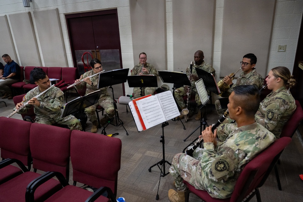 A group of seven military personnel in camouflage uniforms are seated in a semicircle performing a woodwind and brass ensemble rehearsal in an indoor room. They are reading sheet music from black music stands and playing various wind instruments. The setting appears to be a rehearsal or practice room with red upholstered chairs arranged around the space.