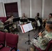 A group of seven military personnel in camouflage uniforms are seated in a semicircle performing a woodwind and brass ensemble rehearsal in an indoor room. They are reading sheet music from black music stands and playing various wind instruments. The setting appears to be a rehearsal or practice room with red upholstered chairs arranged around the space.