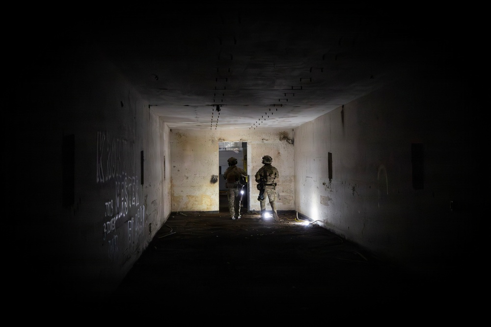 Two armed and helmeted personnel in military/tactical gear are conducting what appears to be a building clearance operation inside a dark, deteriorated corridor. They are positioned at a doorway with tactical flashlights illuminating the space. The structure shows signs of abandonment, with graffiti on the walls and general decay throughout.