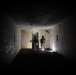 Two armed and helmeted personnel in military/tactical gear are conducting what appears to be a building clearance operation inside a dark, deteriorated corridor. They are positioned at a doorway with tactical flashlights illuminating the space. The structure shows signs of abandonment, with graffiti on the walls and general decay throughout.