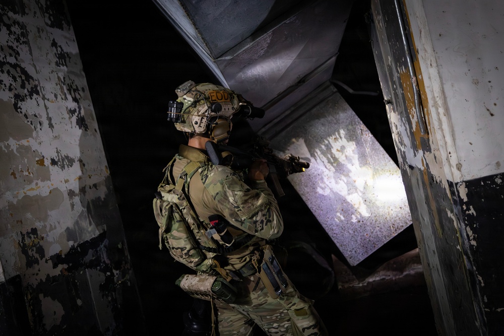 A fully equipped military or law enforcement operator wearing multicam camouflage and a tactical helmet with 'EOD' markings is conducting a building clearance operation in a dilapidated structure. The operator is positioned at a doorway or threshold, aiming a weapon with an attached weapon light illuminating the space ahead. The environment shows heavily deteriorated walls with peeling paint, suggesting an abandoned or damaged building.