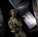 A fully equipped military or law enforcement operator wearing multicam camouflage and a tactical helmet with 'EOD' markings is conducting a building clearance operation in a dilapidated structure. The operator is positioned at a doorway or threshold, aiming a weapon with an attached weapon light illuminating the space ahead. The environment shows heavily deteriorated walls with peeling paint, suggesting an abandoned or damaged building.