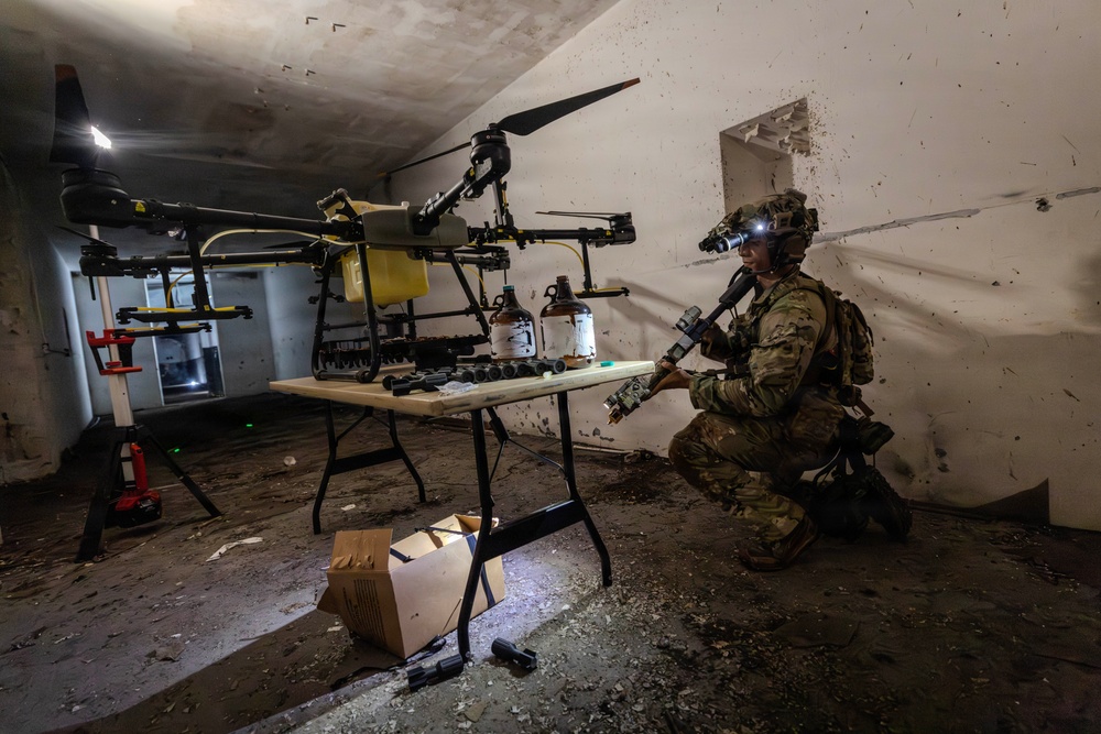 A soldier in full combat gear with night-vision equipment kneels inside a damaged interior space, examining what appears to be a drone component or device. The room contains a large multi-rotor drone (likely an agricultural or modified UAV) on a stand, a folding table with various containers and components, and scattered debris. The scene appears to depict a military training exercise or simulated discovery of an improvised drone facility.