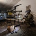 A soldier in full combat gear with night-vision equipment kneels inside a damaged interior space, examining what appears to be a drone component or device. The room contains a large multi-rotor drone (likely an agricultural or modified UAV) on a stand, a folding table with various containers and components, and scattered debris. The scene appears to depict a military training exercise or simulated discovery of an improvised drone facility.