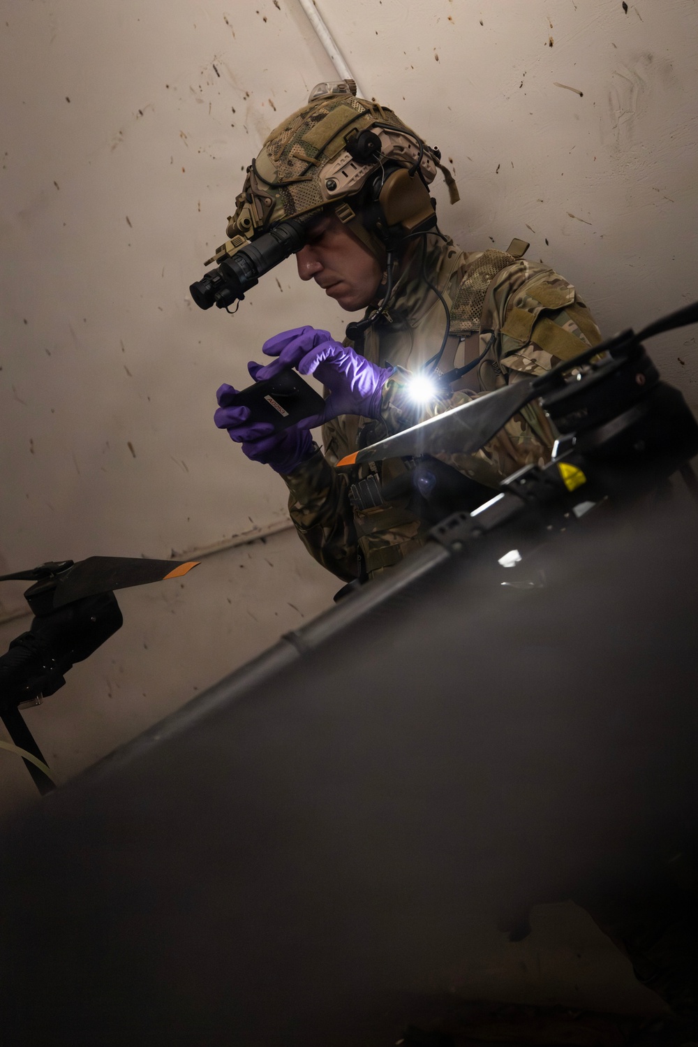 A military or law enforcement operator in full tactical gear is examining what appears to be a drone or UAS (Unmanned Aerial System) in a dimly lit interior space. The individual is wearing multicam camouflage, a tactical helmet with a mounted device (likely a night vision monocular), and purple nitrile gloves, and is using a handheld device with an active flashlight to inspect the equipment. The scene suggests a forensic examination, exploitation, or technical assessment of a captured/recovered drone.