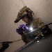 A military or law enforcement operator in full tactical gear is examining what appears to be a drone or UAS (Unmanned Aerial System) in a dimly lit interior space. The individual is wearing multicam camouflage, a tactical helmet with a mounted device (likely a night vision monocular), and purple nitrile gloves, and is using a handheld device with an active flashlight to inspect the equipment. The scene suggests a forensic examination, exploitation, or technical assessment of a captured/recovered drone.