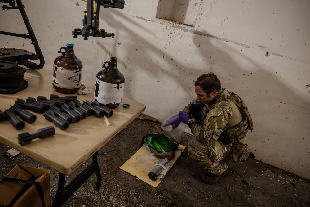 A soldier in multicam/OCP camouflage uniform and tactical vest is kneeling on the ground wearing purple nitrile gloves, examining or handling items from a bag. A folding table nearby holds multiple dark-colored cylindrical components and two large amber glass jugs with partially removed labels. The setting appears to be a damaged or weathered interior space, consistent with a training or field exploitation scenario.