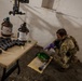 A soldier in multicam/OCP camouflage uniform and tactical vest is kneeling on the ground wearing purple nitrile gloves, examining or handling items from a bag. A folding table nearby holds multiple dark-colored cylindrical components and two large amber glass jugs with partially removed labels. The setting appears to be a damaged or weathered interior space, consistent with a training or field exploitation scenario.