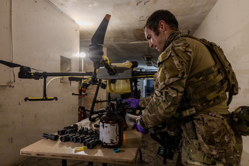 A person in military camouflage uniform and tactical vest is working at a table in what appears to be a basement or bunker environment. They are wearing purple nitrile gloves and handling containers near a large multi-rotor drone (appears to be an agricultural-style drone) positioned behind them. Various cylindrical components and containers are laid out on the work table.