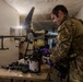 A person in military camouflage uniform and tactical vest is working at a table in what appears to be a basement or bunker environment. They are wearing purple nitrile gloves and handling containers near a large multi-rotor drone (appears to be an agricultural-style drone) positioned behind them. Various cylindrical components and containers are laid out on the work table.