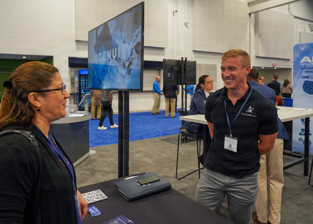 A trade show or conference exhibition floor where a male booth representative wearing a 'SpaceWERX' branded polo shirt is engaged in conversation with a female attendee. The booth features a monitor displaying the word 'INGENUITY' and has a QR code visible on the table. The background shows additional exhibitors and attendees in a large convention hall with blue carpeting.