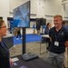 A trade show or conference exhibition floor where a male booth representative wearing a 'SpaceWERX' branded polo shirt is engaged in conversation with a female attendee. The booth features a monitor displaying the word 'INGENUITY' and has a QR code visible on the table. The background shows additional exhibitors and attendees in a large convention hall with blue carpeting.