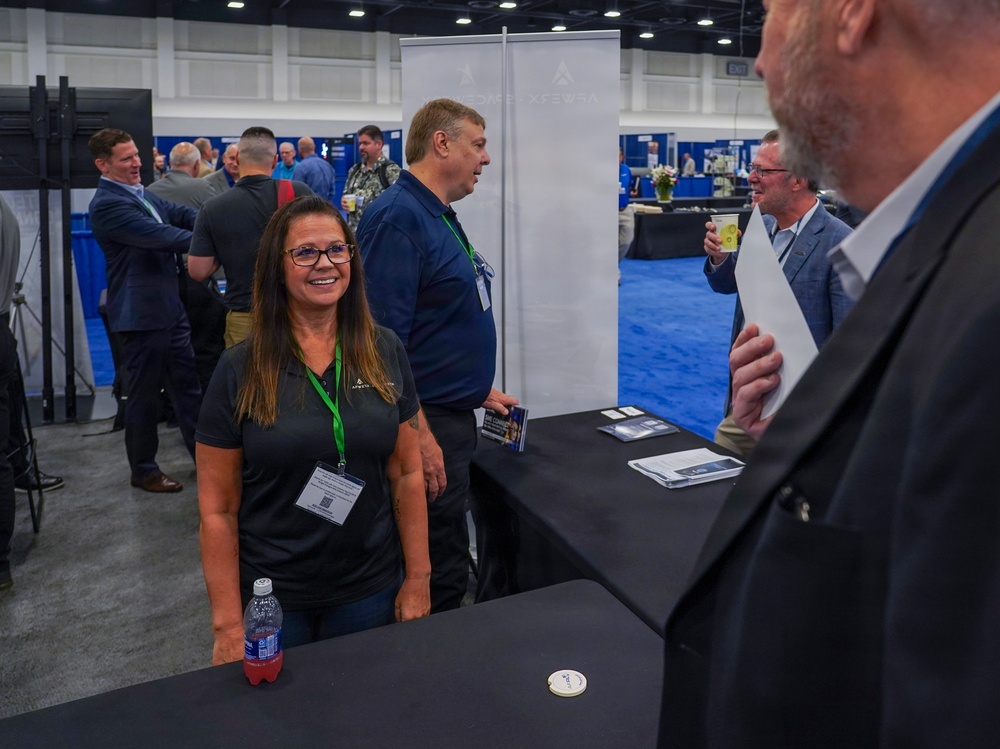 A professional trade show or conference exhibition floor with multiple vendor booths and attendees networking. A woman in a dark polo shirt with a green lanyard badge smiles at the foreground booth, accompanied by a male colleague behind her. The event appears to be a business or defense/aerospace industry conference based on the branding visible on the banner.
