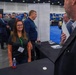 A professional trade show or conference exhibition floor with multiple vendor booths and attendees networking. A woman in a dark polo shirt with a green lanyard badge smiles at the foreground booth, accompanied by a male colleague behind her. The event appears to be a business or defense/aerospace industry conference based on the branding visible on the banner.