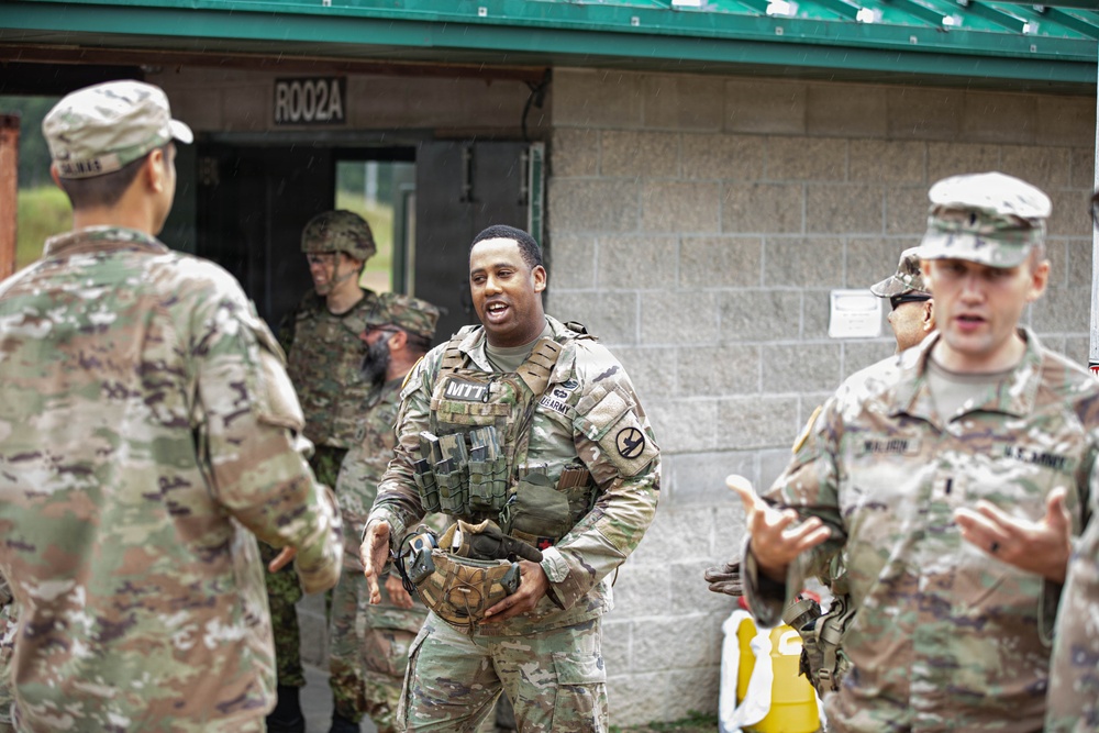 A group of U.S. Army soldiers in OCP (Operational Camouflage Pattern) uniforms are gathered outside a concrete block building marked 'R002A' with a green roof. The central figure, wearing a tactical vest marked 'MTT' (Mobile Training Team), appears to be engaged in conversation with fellow soldiers. The setting appears to be a military training facility, with rain visible in the background.