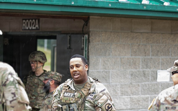 A group of U.S. Army soldiers in OCP (Operational Camouflage Pattern) uniforms are gathered outside a concrete block building marked 'R002A' with a green roof. The central figure, wearing a tactical vest marked 'MTT' (Mobile Training Team), appears to be engaged in conversation with fellow soldiers. The setting appears to be a military training facility, with rain visible in the background.