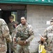 A group of U.S. Army soldiers in OCP (Operational Camouflage Pattern) uniforms are gathered outside a concrete block building marked 'R002A' with a green roof. The central figure, wearing a tactical vest marked 'MTT' (Mobile Training Team), appears to be engaged in conversation with fellow soldiers. The setting appears to be a military training facility, with rain visible in the background.
