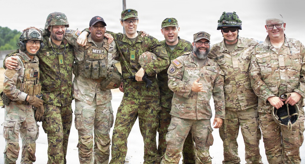 A group of eight military personnel in various camouflage uniforms pose together in a casual, jovial manner, smiling and laughing for the camera. The group appears to include both U.S. Army soldiers and what appear to be allied nation military members wearing different camouflage patterns. The setting appears to be an outdoor military installation or training area.