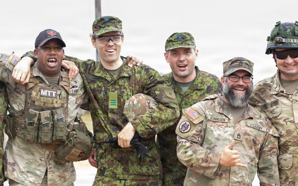 A group of eight military personnel in various camouflage uniforms pose together in a casual, jovial manner, smiling and laughing for the camera. The group appears to include both U.S. Army soldiers and what appear to be allied nation military members wearing different camouflage patterns. The setting appears to be an outdoor military installation or training area.