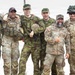 A group of eight military personnel in various camouflage uniforms pose together in a casual, jovial manner, smiling and laughing for the camera. The group appears to include both U.S. Army soldiers and what appear to be allied nation military members wearing different camouflage patterns. The setting appears to be an outdoor military installation or training area.