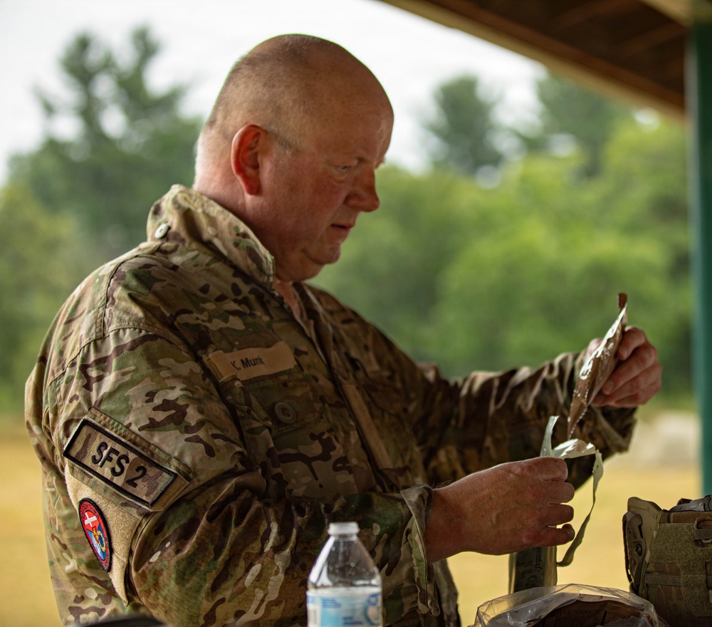 A middle-aged male military personnel wearing MultiCam/OCP camouflage uniform is handling what appears to be equipment or a pouch under an outdoor covered structure. His uniform displays a name tape reading 'K. Munt', a patch labeled 'SFS 2', and a circular unit/organizational patch on the left sleeve. The setting appears to be a field training or operational environment with trees visible in the background.