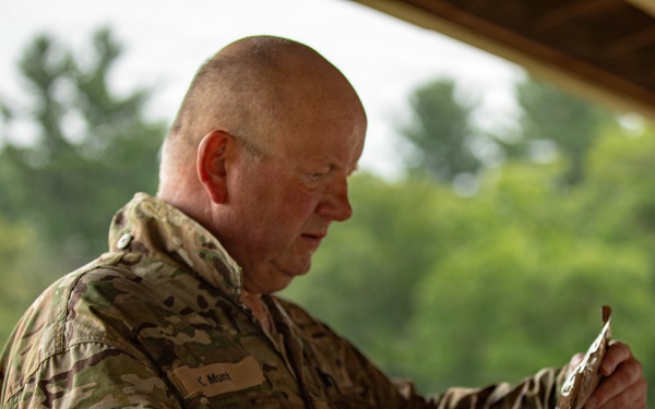 A middle-aged male military personnel wearing MultiCam/OCP camouflage uniform is handling what appears to be equipment or a pouch under an outdoor covered structure. His uniform displays a name tape reading 'K. Munt', a patch labeled 'SFS 2', and a circular unit/organizational patch on the left sleeve. The setting appears to be a field training or operational environment with trees visible in the background.
