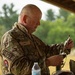 A middle-aged male military personnel wearing MultiCam/OCP camouflage uniform is handling what appears to be equipment or a pouch under an outdoor covered structure. His uniform displays a name tape reading 'K. Munt', a patch labeled 'SFS 2', and a circular unit/organizational patch on the left sleeve. The setting appears to be a field training or operational environment with trees visible in the background.