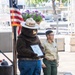 The image shows Smokey Bear mascot costume standing beside a uniformed forest service ranger at what appears to be an outdoor public event. Smokey Bear is holding a framed certificate or award, wearing his iconic white ranger hat labeled 'SMOKEY', blue jeans with a 'SMOKEY' belt buckle, and brown bear feet. An American flag is visible to the left, and a hanging basket of purple flowers is in the background.